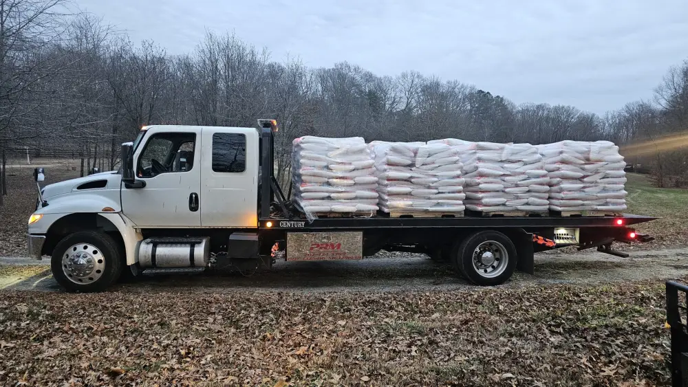 PRM flatbed truck loaded with pallets of shrink-wrapped bagged material on a gravel drive.